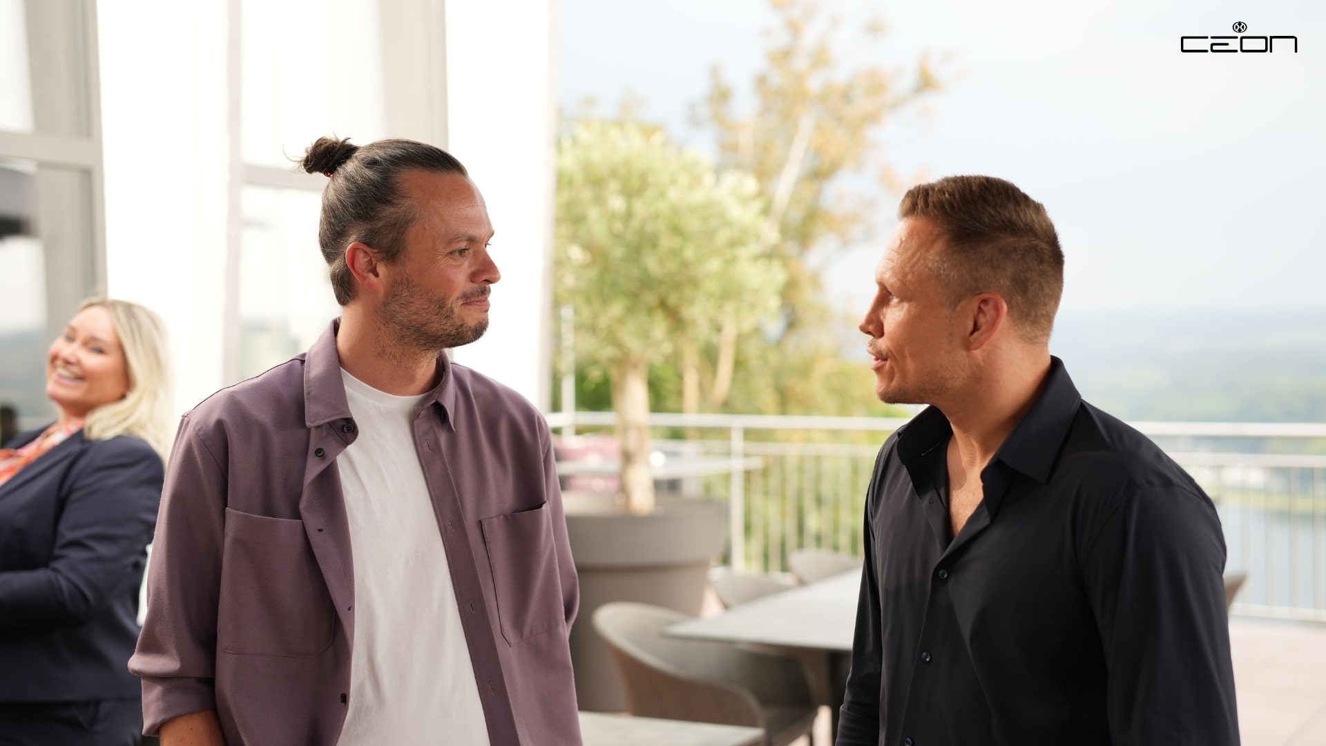Zwei Männer unterhalten sich lächelnd im Freien, Mann links trägt lila Hemd, rechts schwarzes Hemd.