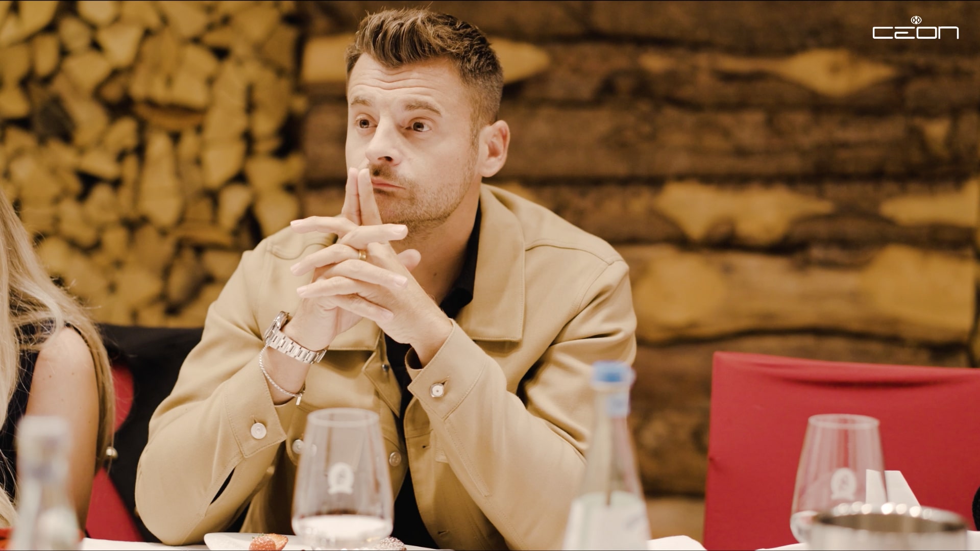 Ein nachdenklicher Mann, trägt eine beige Jacke, sitzt an einem Tisch mit Holzwand im Hintergrund.