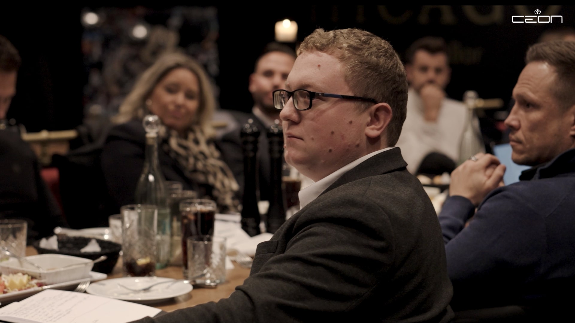 Eine Person mit Brille sitzt, neutraler Ausdruck, in einem formellen Meeting, umgeben von Personen.