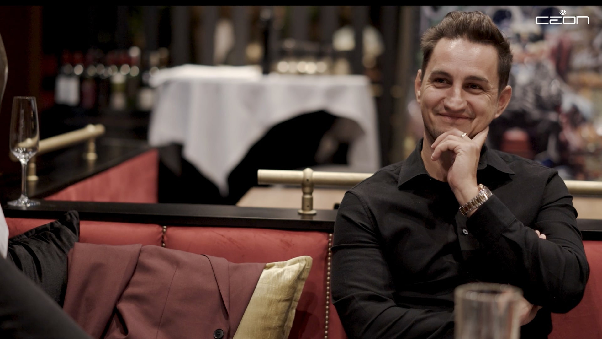 Ein lächelnder Mann in schwarzem Hemd sitzt auf rotem Sofa in einem eleganten Restaurant.