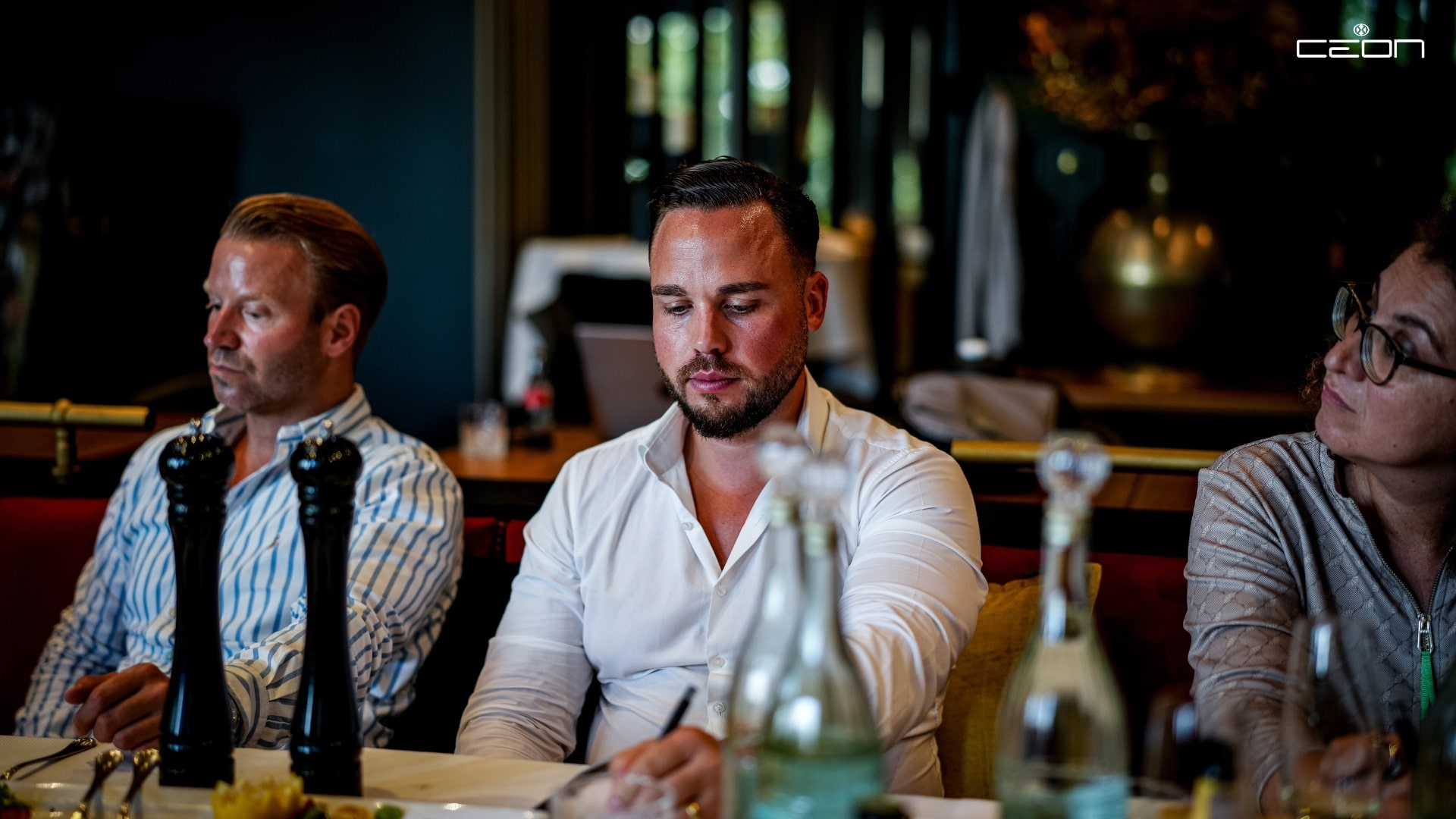 Drei Personen am Tisch, nachdenklich, elegante Kleidung, Flaschen und Pfeffermühlen im Vordergrund.