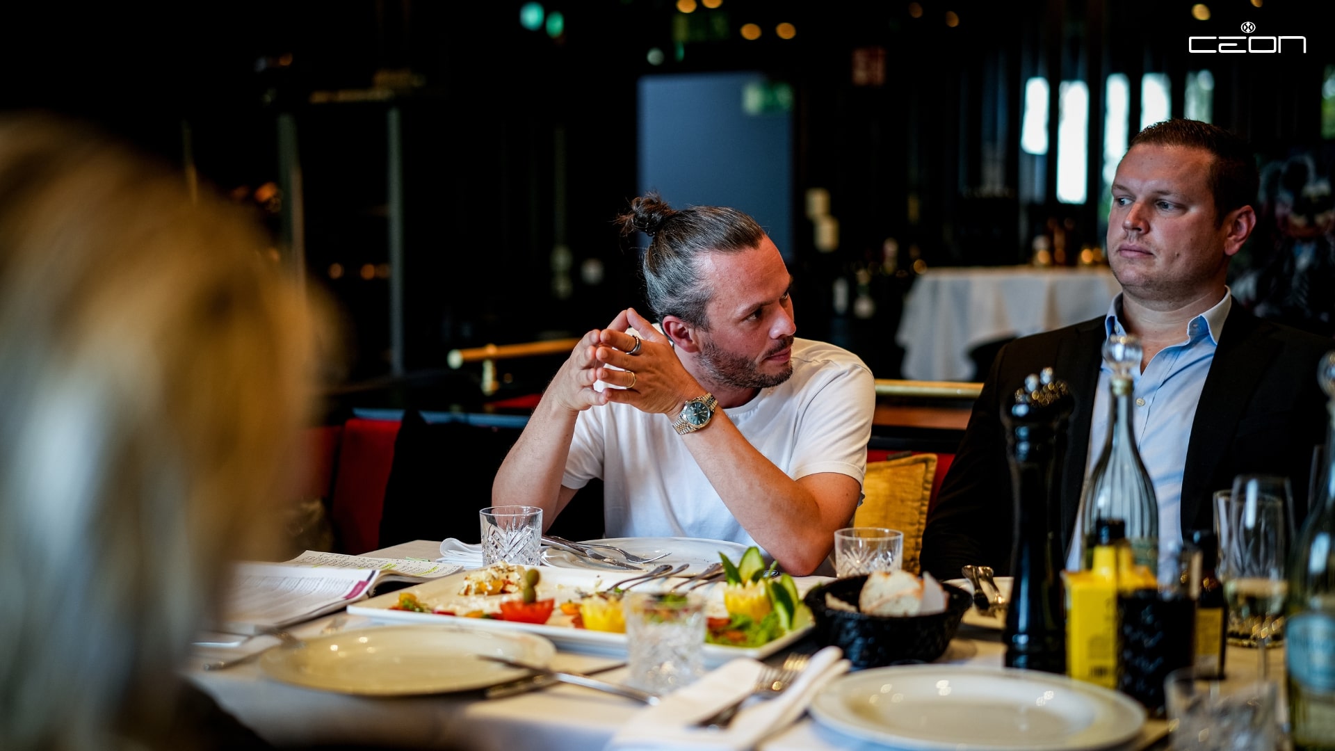 Ein Mann mit Man Bun spricht mit einem ernst aussehenden Mann im Restaurant. Bunte Tischdekoration.