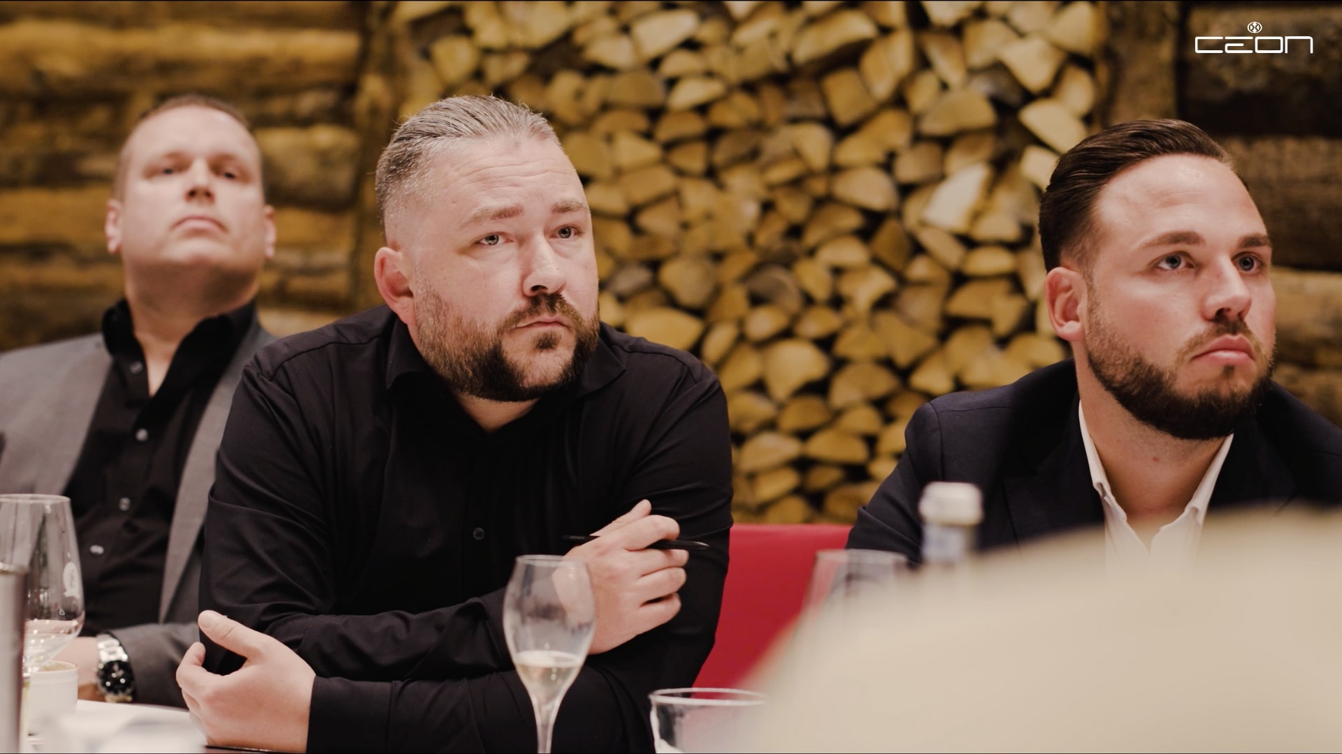 Drei Männer in Anzügen schauen konzentriert. Hintergrund mit gestapeltem Holz.