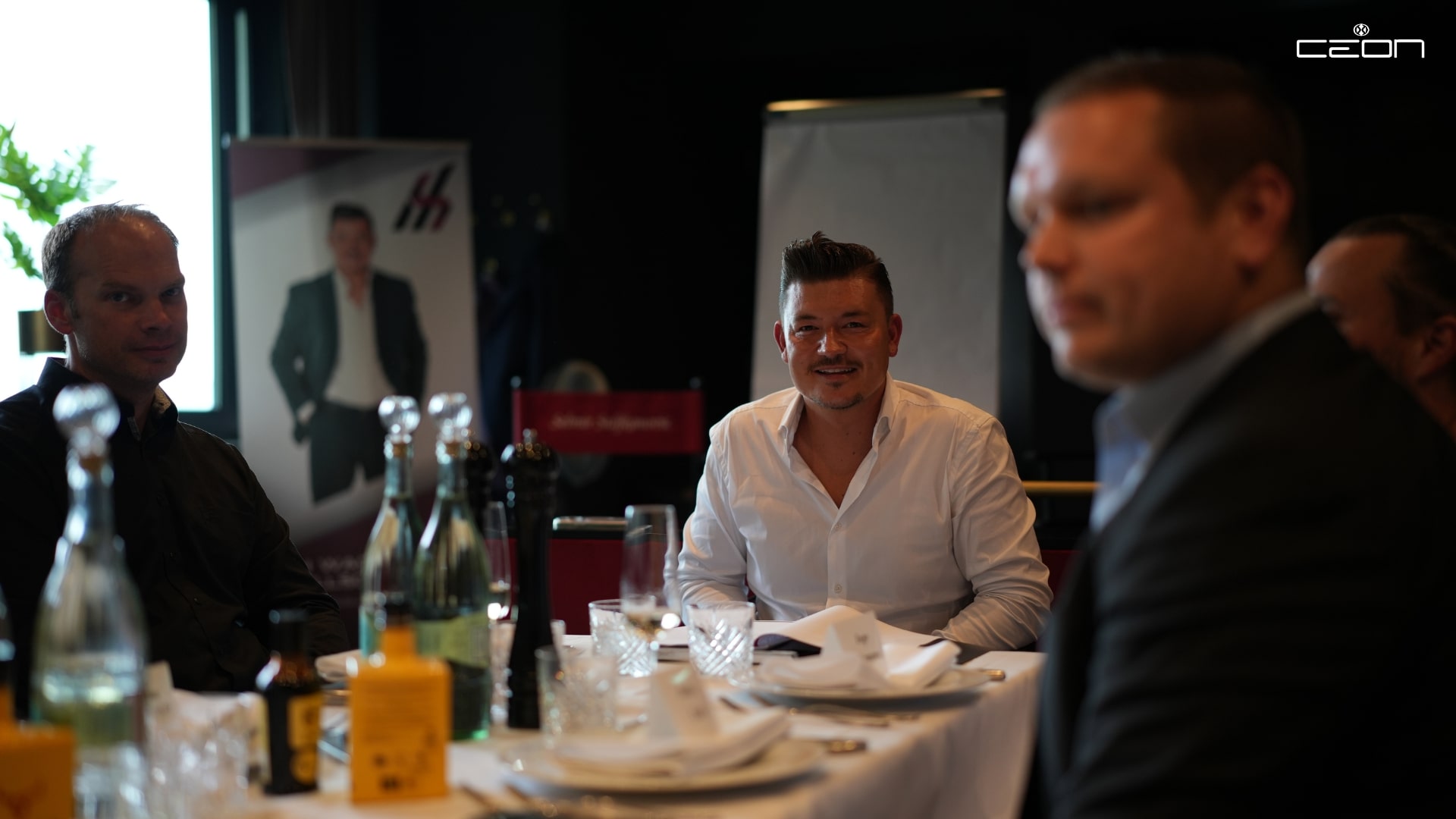 Vier Männer sitzen an einem Tisch, einer lächelt. Weißes Hemd, Wasserflaschen und Gläser.