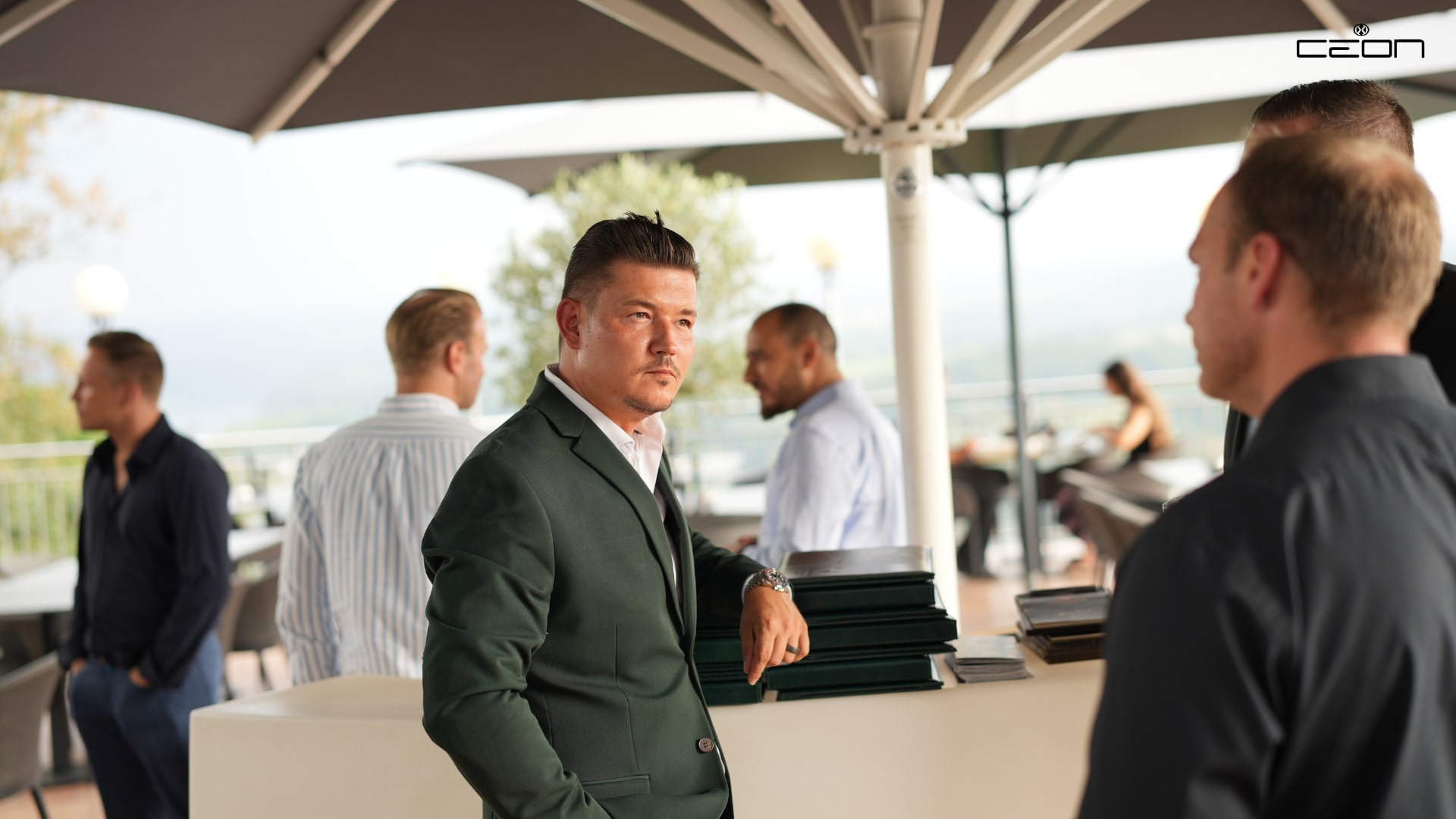 Ein Mann in grünem Anzug mit ernstem Blick steht an einer Bar im Freien, umgeben von Personen.